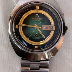 May include: Vintage Ricoh automatic watch with a stainless steel band. The watch face is green and gold with the day and date displayed. The watch is water resistant and has 21 jewels.