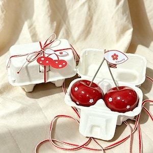 May include: Two glossy red cherry-shaped objects with small white flower patterns, each featuring a thin metal stem and a paper leaf tag labeled Cherry, presented inside a white molded pulp egg carton tied with red and white twine.