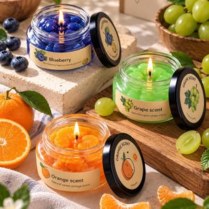 May include: Three glass jar candles with black lids, featuring blueberry, grape, and orange scents. Each candle contains wax shaped like fruit pieces and has a label with the corresponding fruit name and scent description. The candles are lit.