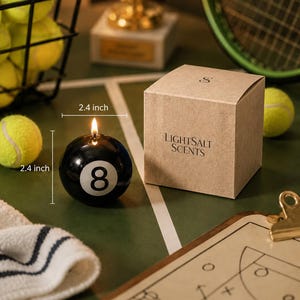 May include: A black spherical candle shaped like an 8-ball with a lit wick, measuring 2.4 inches in diameter. Next to it is a square tan cardboard box labeled LIGHTSALT SCENTS with a small S logo on top, set against a sports-themed background.