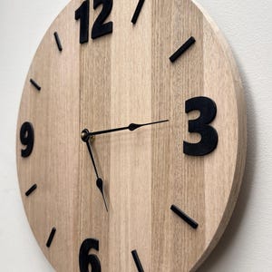 Peut inclure: Une horloge murale ronde en bois avec une finition marron clair. L'horloge comporte des chiffres et des marqueurs de minutes noirs. Les aiguilles de l'horloge sont noires. L'horloge mesure environ 30 cm de diamètre.