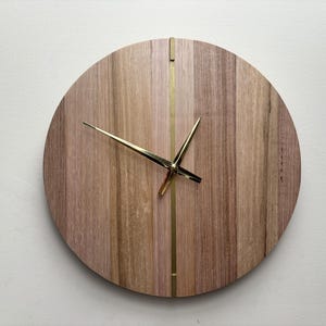 Peut inclure: Une horloge murale ronde en bois avec un fini grain de bois naturel. Le cadran de l'horloge présente des panneaux de bois verticaux dans des tons de marron et de beige, avec une fine bande dorée au centre. Les aiguilles dorées indiquent l'heure.
