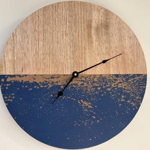 Peut inclure: Une horloge murale ronde avec un design bicolore. La moitié supérieure est en bois naturel, et la moitié inférieure est bleu marine avec des éclaboussures dorées. Les aiguilles noires indiquent l'heure.