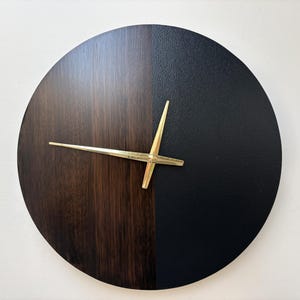 Puede incluir: Un reloj de pared redondo con un diseño bicolor. La mitad de la esfera es de madera oscura, la otra mitad es negra. Las manecillas del reloj son doradas. Este reloj moderno es un elemento decorativo para el hogar.
