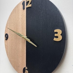Peut inclure: Une horloge murale ronde bicolore. La moitié du cadran est en bois naturel, l'autre moitié est noire. Des chiffres dorés indiquent les positions 12, 3, 6 et 9 heures. Les aiguilles de l'horloge sont dorées.