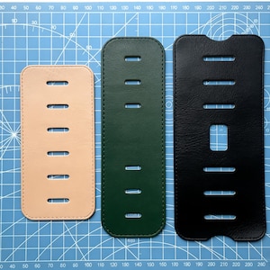 Peut inclure: Trois étuis de téléphone en cuir beige, vert et noir, chacun avec des découpes horizontales. Les étuis sont rectangulaires avec des coins arrondis, disposés sur un tapis de coupe quadrillé bleu. L'étui noir a une découpe centrale plus grande. Mesures en cm.