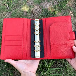 Peut inclure: Un classeur en cuir rouge ouvert avec des anneaux dorés et une colonne vertébrale noire. Le classeur comporte plusieurs poches et une fermeture à bouton-pression. Le classeur est maintenu ouvert, révélant l'intérieur. L'arrière-plan est de l'herbe verte.