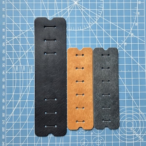 May include: Three leather straps in black, tan, and gray, each with a series of evenly spaced holes and a notched end. The straps are arranged vertically against a blue cutting mat, showcasing their different colors and textures.