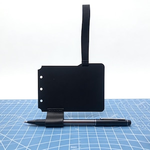 Op de afbeelding: Een zwarte leren pennenhouder met een bijpassend label en een zilveren en zwarte pen. De pennenhouder is bevestigd aan een zwart label met drie gaten en een zwarte band. De pen ligt op een blauwe snijmat.