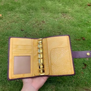 May include: A purple leather binder with a yellow interior featuring a six-ring gold metal mechanism. The left side includes multiple card slots and a clear window pocket, while the right side has a large slip pocket and a snap button closure.