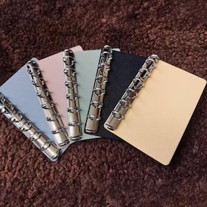 May include: A collection of five small notebooks with silver metal ring binders. The notebooks have covers in pastel colors: light blue, pink, green, navy, and cream. The notebooks are arranged on a brown, textured surface.