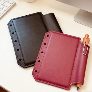 May include: Two leather planner covers, one dark brown and one burgundy, are displayed on a white surface. The burgundy cover has a pen holder with an orange pen. Both covers have a series of evenly spaced holes along the left edge.