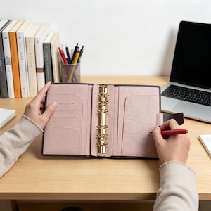 May include: Open pink leather binder with gold-colored rings. The binder is open, revealing card slots and a pen holder. A red pen is held near the right side of the binder. A laptop and books are in the background.