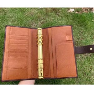May include: A tan leather personal organizer featuring a gold-toned six-ring metal binder mechanism in the center. The interior includes multiple vertical card slots on the left side, a full-length pocket, and a pen loop on the right side.