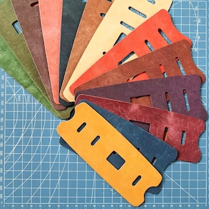 May include: A collection of colorful leather pieces in various shades, including orange, green, and brown. Each piece has a unique shape with cut-out sections and a textured surface. The items are arranged on a blue cutting mat.