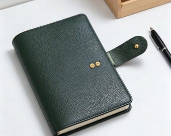 May include: A dark green leather journal with a textured finish. The journal features a flap closure with a gold press stud and two gold rivets. A black pen lies on the white surface beside the journal. A wooden desk organiser is in the background.