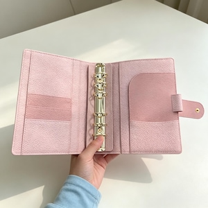 May include: Open, light pink binder with a gold-colored ring mechanism. The binder has multiple pockets and a snap closure. The interior is a lighter shade of pink than the exterior. The binder is held open, displaying its features.