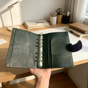 Puede incluir: Carpeta de cuero verde oscuro abierta, con anillos plateados y un cierre morado. La carpeta tiene múltiples bolsillos y una superficie texturizada. La carpeta está abierta, revelando el diseño interior.