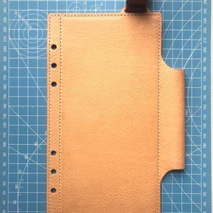 以下が含まれることがあります： 茶色のストラップが付いた、明るいタン色の革製プランナーカバー。 カバーの左側には一連の穴があり、右側には湾曲したポケットがあります。 カバーは青いグリッド上にあります。