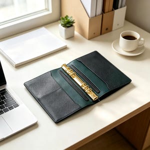 May include: An open six-ring binder featuring a textured black exterior and a dark green interior. The center mechanism is made of polished gold-tone metal, designed to hold loose-leaf paper. The binder includes two interior pockets for storage.