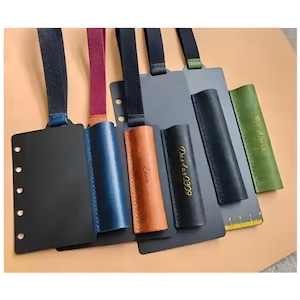 Puede incluir: Una colección de portalápices y cubiertas de cuadernos de cuero en varios colores, incluyendo negro, azul, marrón y verde. Algunos portadores tienen texto, y todos tienen correas. Las cubiertas de los cuadernos tienen una fila de agujeros para encuadernar.