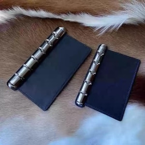 Op de afbeelding: Twee kleine, donkerblauwe leren notitieboekjes met zilveren metalen ringbanden. De notitieboekjes zijn rechthoekig en liggen op een bruin en wit vachtachtig oppervlak. De ringbanden zijn cilindrisch.