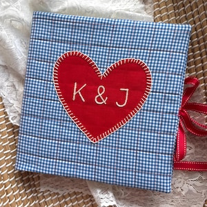 May include: A square fabric album cover featuring a blue and white gingham pattern with quilted stitching. A large red fabric heart is appliqued in the centre with white blanket stitching and the embroidered white letters K & J. Red ribbons are attached.