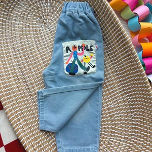 May include: Pale blue denim trousers with an elasticated waistband. The back pocket displays a colourful graphic with the word "APPLE" and cartoon characters. The trousers are folded on a woven basket.