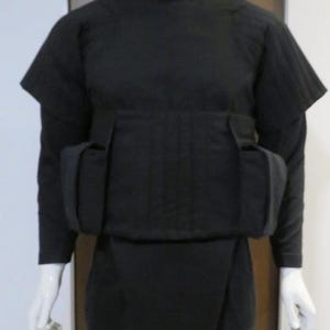 May include: A black costume displayed on a mannequin. The outfit includes a high-collared top with short sleeves, a chest piece with two pockets, and form-fitting pants. The overall design is sleek and monochromatic.