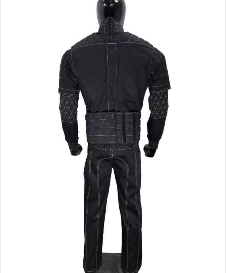 May include: A black tactical-style jumpsuit featuring a high collar, long sleeves with white geometric grid stitching on the forearms, a padded waist belt, and white contrast stitching throughout the garment, displayed on a mannequin.