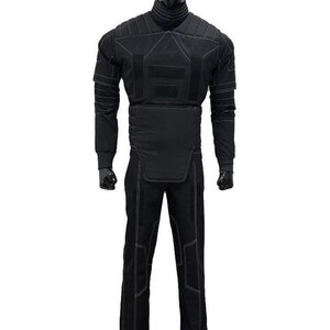May include: A black tactical-style costume set featuring a long-sleeved top with a high neck, a padded chest and waist panel, and matching trousers. The entire outfit is accented with visible white stitching and rectangular fabric patches.