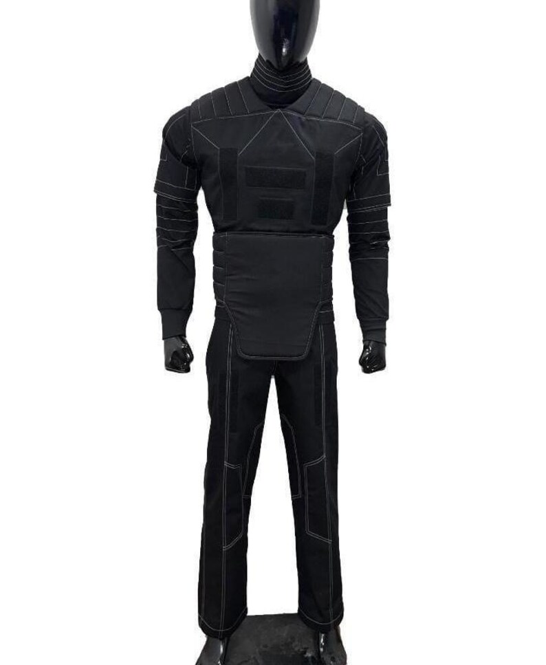 May include: A black tactical-style costume set featuring a long-sleeved top with a high neck, a padded chest and waist panel, and matching trousers. The entire outfit is accented with visible white stitching and rectangular fabric patches.