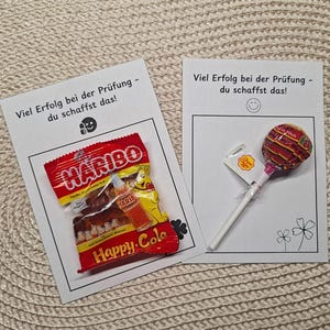 Può includere: Due biglietti bianchi con la scritta "In bocca al lupo per il tuo esame - ce la puoi fare!" e una faccina sorridente. Un biglietto ha un sacchetto di caramelle Haribo Happy-Cola, l'altro un lecca lecca Chupa Chups.