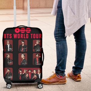 Op de afbeelding: Zwarte rolkoffer met de tekst "BTS WORLD TOUR" in het rood en afbeeldingen van de bandleden. De koffer heeft een zwarte handgreep en vier wielen.