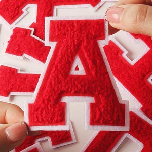 Op de afbeelding: Rode vilt letters met witte randen, verspreid over een wit oppervlak. De letter "A" staat centraal, vastgehouden door een hand. De afbeelding bevat de tekst "PresentsNest design". Ideaal voor knutselen en personaliseren.