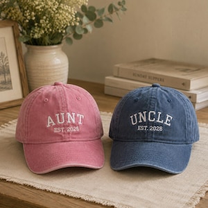 May include: A pink baseball cap with white embroidered text reading AUNT EST. 2026 and a blue baseball cap with white embroidered text reading UNCLE EST. 2026. Both caps feature a structured crown, a curved brim, and a washed fabric finish.