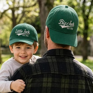 Puede incluir: Dos gorras de béisbol verde bosque con texto bordado en blanco. La gorra más pequeña muestra 'Little Jack' con 'EST. 2021' debajo, mientras que la más grande muestra 'Big Jack' con 'EST. 1980' debajo, ambas con un gráfico de swoosh blanco.