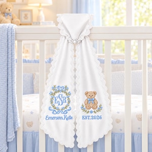 Op de afbeelding: Een witte stoffen babydeken met geschulpte kanten randen, voorzien van blauw borduurwerk van het monogram ESR, de naam Emerson Kate, een bruine teddybeer met een blauwe strik en de tekst EST. 2026, gedrapeerd over een witte ledikantrand.