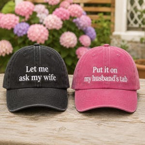 May include: A black baseball cap with the white embroidered text Let me ask my wife and a pink baseball cap with the white embroidered text Put it on my husband's tab. Both caps feature a curved brim and a structured crown design.