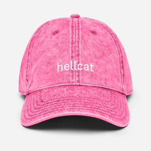 Op de afbeelding: Roze baseballpet met het woord "hellcat" in witte letters. De pet heeft een gebogen klep en een verweerde, vintage uitstraling.