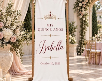 May include: A vertical off-white fabric banner suspended from a wooden frame. The banner features a gold crown graphic, the text "MIS QUINCE AÑOS", the name "Isabella" in a dark red script font, and the date "October 10, 2026" in dark red text.