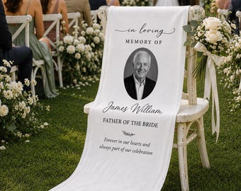 May include: A white memorial sign draped over a wooden chair, featuring the text "in loving MEMORY OF", a black and white oval portrait of a man, the name "James William", "FATHER OF THE BRIDE", and the phrase "Forever in our hearts and always part of our celebration".