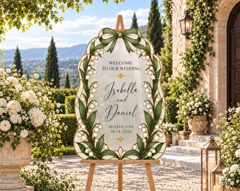 Könnte beinhalten: Ein Hochzeits-Willkommensschild auf einer Holzstaffelei mit gewelltem Rand, einer grünen Schleife oben und Maiglöckchen-Illustrationen. Der Text lautet: Welcome to our wedding, Isabella and Daniel, Austin, USA, 14.10.2026.