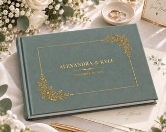 May include: A sage green guest book with gold foil lettering that reads ALEXANDRA & KYLE and SEPTEMBER 19, 2026. The cover features a rectangular gold frame with decorative floral corner accents and a gold pen resting on the surface.