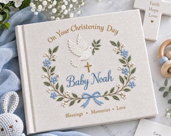 Könnte beinhalten: Ein cremefarbenes Hardcover-Buch mit gestickten Details, darunter eine weiße Taube, ein goldenes Kreuz, blaue Blumen und grüne Blätter. Der Text lautet On Your Christening Day, Baby Noah sowie Blessings, Memories, Love in Gold und Blau.