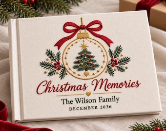 May include: A white fabric-covered photo album featuring embroidered Christmas decorations including a red bow, a gold ornament with a green tree, and red berries. The text reads Christmas Memories, The Wilson Family, and DECEMBER 2026.