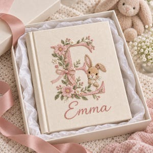 May include: A cream-colored fabric-covered book featuring an embroidered large letter E decorated with pink flowers, green leaves, a small bow, and a brown bunny. The name Emma is embroidered in pink cursive script below the letter E.