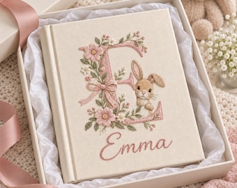 Könnte beinhalten: Ein cremefarbenes, stoffbezogenes Buch mit einem gestickten großen Buchstaben E, verziert mit rosa Blumen, grünen Blättern, einer kleinen Schleife und einem braunen Hasen. Der Name Emma ist in rosa Schreibschrift unter dem E gestickt.