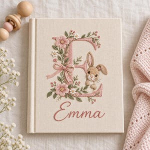 May include: A cream-colored fabric-covered book featuring an embroidered large letter E decorated with pink flowers, green leaves, and a small brown rabbit. The name Emma is embroidered in pink cursive script below the letter E on the front cover.