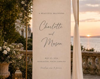 Könnte beinhalten: Ein hohes, rechteckiges Hochzeitsschild aus Stoff, das an einem goldfarbenen Metallrahmen hängt, mit dem Text A BEAUTIFUL BEGINNING, Charlotte and Mason, MAY 22, 2026, CHARLESTON, SOUTH CAROLINA und timeless vows & forever in schwarzer Schrift.